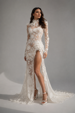 Load image into Gallery viewer, Woman wearing the Elysian Bloom Couture Gown featuring floral embroidered lace, illusion mesh, high neckline, sheer long sleeves, three-dimensional floral appliqués, and a dramatic thigh-high slit.