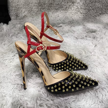 Load image into Gallery viewer, Black pointed-toe stiletto pumps with gold stud embellishments, red patent ankle straps, and sculptural metallic heels displayed in a luxury fashion setting.