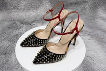 Load image into Gallery viewer, Black pointed-toe stiletto pumps with gold stud embellishments, red patent ankle straps, and sculptural metallic heels displayed in a luxury fashion setting.