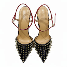 Load image into Gallery viewer, Black pointed-toe stiletto pumps with gold stud embellishments, red patent ankle straps, and sculptural metallic heels displayed in a luxury fashion setting.