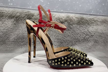 Load image into Gallery viewer, Black pointed-toe stiletto pumps with gold stud embellishments, red patent ankle straps, and sculptural metallic heels displayed in a luxury fashion setting.